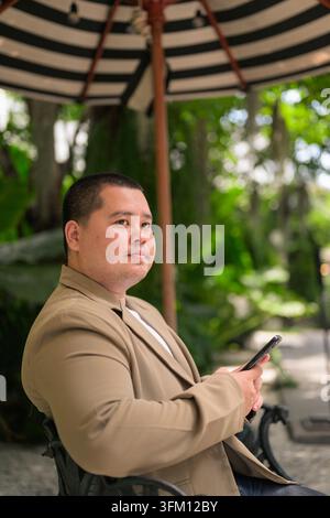 Un giovane uomo d'affari asiatico della regione di Isan, seduto all'aperto e attento a guardare mentre tiene uno smartphone sotto un stri Foto Stock