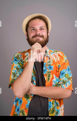 Ritratto in studio di un giovane barbuto sorridente che indossa una colorata camicia arancione tropicale e un cappello di paglia, tenendo il mento premuroso. Concetto per il piano Foto Stock