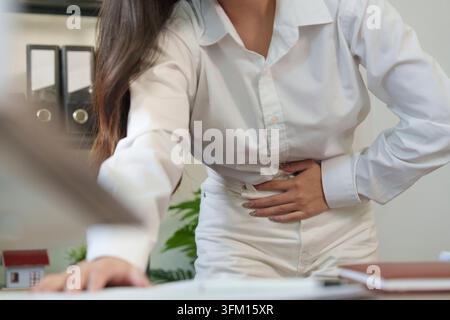Giovane donna asiatica che soffre di dolori allo stomaco mentre lavora in ufficio, illustrando il disagio addominale e potenziali problemi digestivi Foto Stock