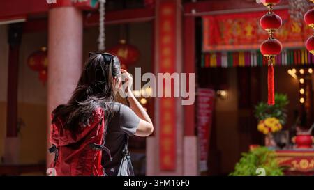 Vista posteriore di una viaggiatrice asiatica con uno zaino rosso che usa la sua fotocamera per scattare una foto di un tempio cinese tradizionale Foto Stock