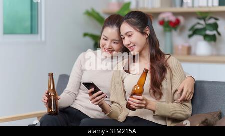 Due amiche asiatiche si rilassano su un divano a casa, una sfoglia un smartphone mentre l'altra tiene una birra, godendosi un tempo libero informale Foto Stock