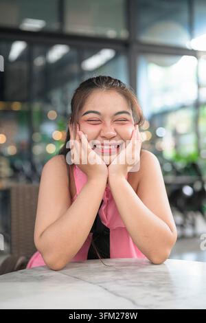Un ritratto verticale all'aperto di una donna Isan thailandese sudorientale sovrappeso sulla ventina. Appoggiando il mento e le guance in entrambe le mani, ride allegramente Foto Stock