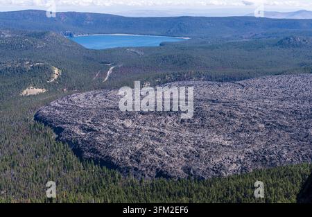 Veduta aerea di un vasto campo di lava basaltica scura che confina con una fitta foresta di pini e le acque blu del lago East in Oregon. Foto Stock