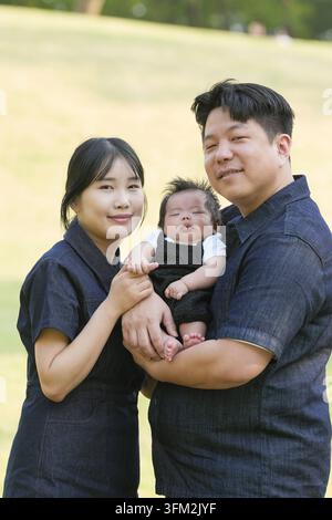 Giovane famiglia che sorride calorosamente all'aperto. Il bambino dorme pacificamente tra le sue braccia. Erba verde e alberi formano uno sfondo morbido. Perfetto per momenti di famiglia o ne Foto Stock