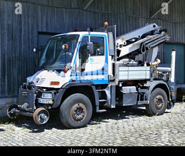 Piccolo camioncino bianco e blu Mercedes-Benz Unimog U 215 con gru ripiegata parcheggiato vicino a un edificio di legno su un pavé Foto Stock