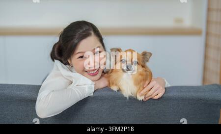 Una donna asiatica felice e il suo piccolo chihuahua si appoggiano sul retro di un divano grigio, sorridendo direttamente alla fotocamera Foto Stock
