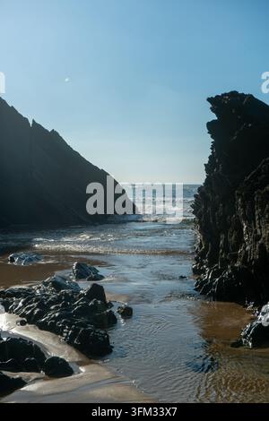 Questa fotografia drammatica mostra una gola stretta scavata in scogliere frastagliate, che conduce a una spiaggia isolata e alla vasta distesa dell'oceano. L'interp Foto Stock