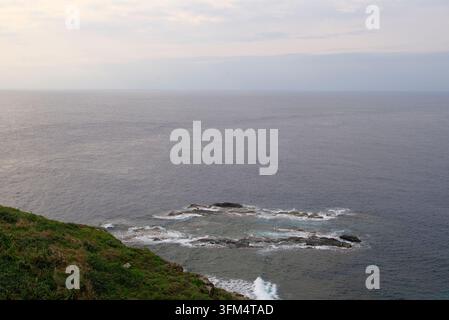 Una vista panoramica ad alta angolazione sull'vastità dell'oceano aperto con onde gentili che si infrangono contro le formazioni rocciose costiere sotto un cielo nuvoloso. Foto Stock