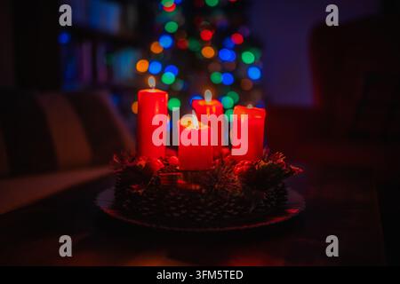 Prima piano di tre candele rosse dell'Avvento accese su una ghirlanda decorata, con luminarie natalizie colorate a quattro luci che creano un bokeh festoso in un interno domestico accogliente Foto Stock