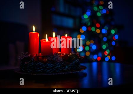Quattro candele dell'Avvento rosse ardenti su una ghirlanda festosa in un interno domestico scuro, con bokeh colorato di albero di Natale e luce ambientale blu fredda nello sfondo Foto Stock