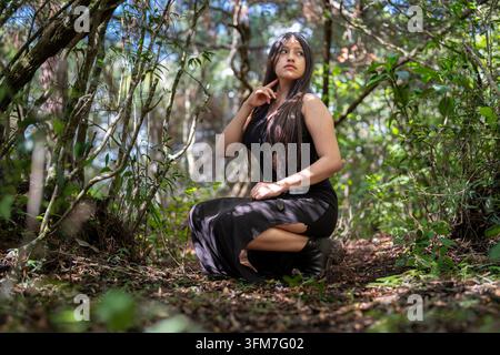 Giovane modella in abito nero accovacciata sul terreno della foresta, che guarda attraverso il fogliame verde. Foto Stock