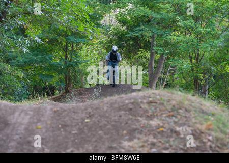 Un pilota con casco integrale guida una moto da cross elettrica sopra i cumuli di terra di una pista da pompaggio circondata da fitti alberi verdi. Foto Stock