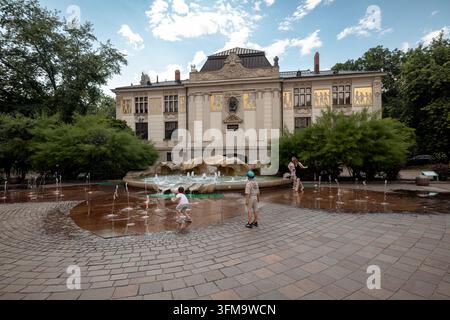 Bambini giocano tra fontane a livello del suolo in Plac Szczepanski con il Palazzo delle Arti sullo sfondo. Foto Stock