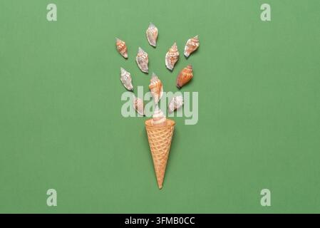 Conchiglie in un cono di waffle per il gelato. Disposizione creativa. Sfondo in carta pastello verde oliva. Vista dall'alto, flat lay. Concetto estivo. Foto Stock