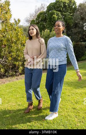 Diverse amiche che camminano attraverso il prato del parco a braccetto indossando maglioni e jeans. Donne, all'aperto, vegetazione, amicizia, maglieria, orologio, luce del giorno Foto Stock