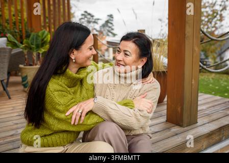 Figlia matura con mamma più grande seduta sul patio di legno, abbracciata. Il concetto del giorno della mamma. Foto Stock