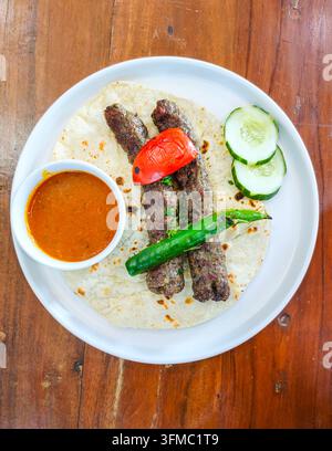 Vista dall'alto a lay piatto di tradizionale kebab Seekh grigliato su pane azzimo con pomodoro grigliato, peperoncino verde, fette di cetriolo e salsa piccante su sfondo bianco Foto Stock