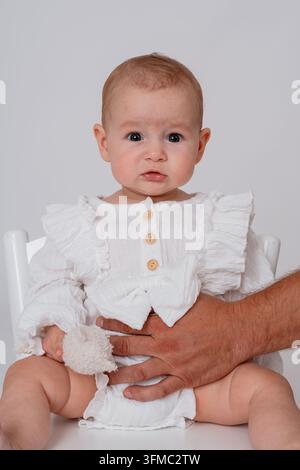 Un bambino sereno e calmo seduto davanti a uno sfondo bianco, con una mano che tiene il tessuto degli abiti del bambino, catturato in un primo piano di famiglia Foto Stock