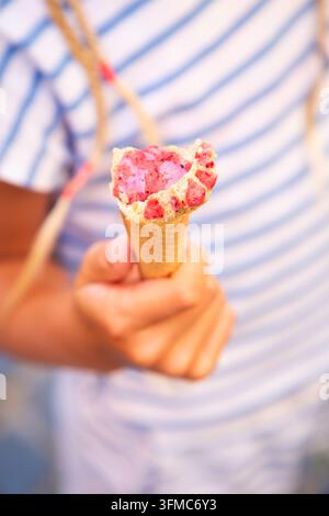 Mano che tiene un cono di gelato alle fragole morso con sfondo sfocato di camicia a strisce, luce calda e soffusa, momento di dessert estivo, vista ravvicinata, casual Foto Stock