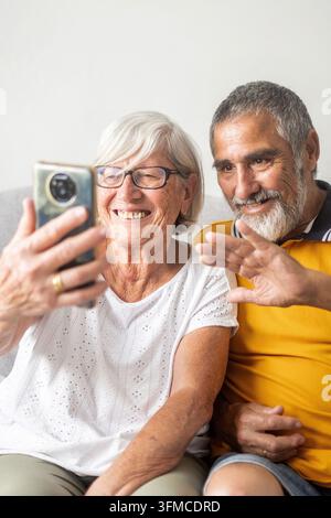 Donna e uomo anziani sorridenti che guardano uno smartphone e salutano durante una videochiamata mentre sono seduti insieme sul divano. Foto Stock