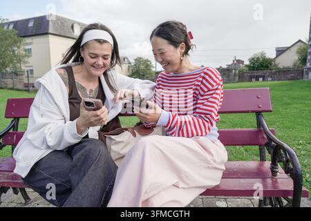 Due donne ridono insieme mentre usano gli smartphone su una panchina del parco Foto Stock