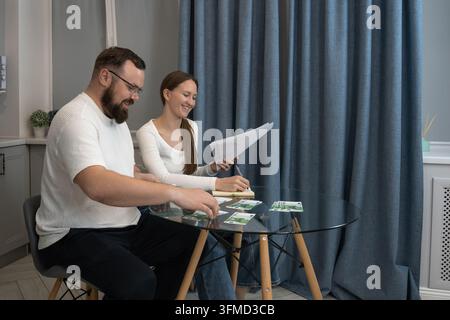 Giovane coppia felice che pianifica il budget domestico, analizza documenti finanziari e calcola le spese con banconote in euro su un tavolo di vetro in un moderno Foto Stock