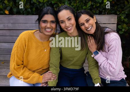 Diverse amiche sedute su una panchina a doghe nel parco, indossano maglioni di lana e un ciondolo smartwatch Foto Stock
