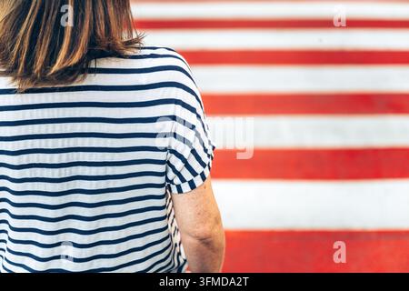 Un primo piano di spalle di una donna in una camicia a righe nere e bianche in piedi davanti a un passaggio pedonale zebrato rosso e bianco dipinto in modo vivace Foto Stock