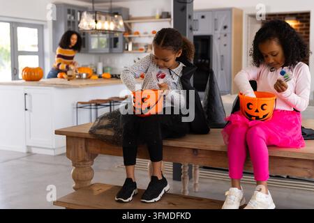 Mamma afroamericana con due figlie sedute in cucina che tengono secchielli di zucca per Halloween Foto Stock