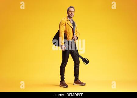 Uomo in giacca gialla e pantaloni di pelle che tiene una chitarra elettrica su uno sfondo giallo Foto Stock