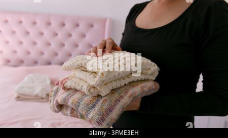 Una donna tiene una pila ordinata di maglieria morbida e appena piegata davanti a uno sfondo sfocato di un letto rosa trapuntato. Questa immagine rappresenta l'organizzazione domestica Foto Stock