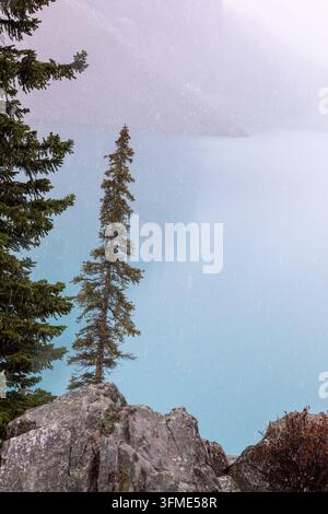 Le acque turchesi del Moraine Lake fanno capolino tra gli alberi sempreverdi e un primo piano roccioso sotto cieli innevati e nuvolosi nel Parco Nazionale di Banff, Canada Foto Stock
