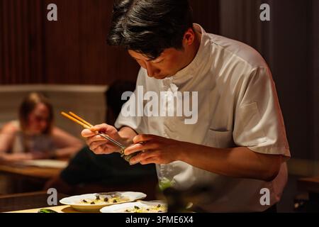 Berlino, Germania – 19 agosto 2025: Uno chef maschile asiatico concentrato prepara abilmente piatti di sushi con le bacchette in un ristorante scarsamente illuminato, mostrando Foto Stock