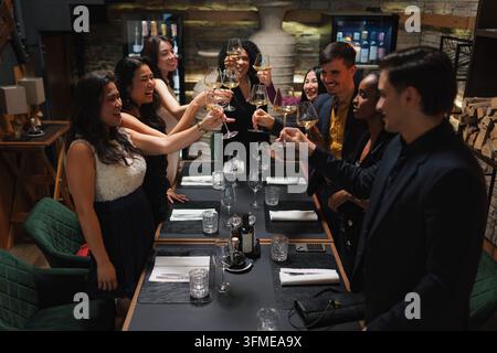 Gruppo di persone diverse, sei amici che passano un bel momento insieme, godendosi la cena in un ristorante moderno di lusso e brindando con il vino. Foto Stock
