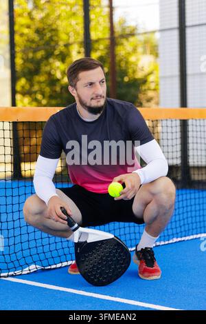 Giocatore di padel seduto accanto alla rete con una racchetta e una palla su un campo al coperto, che illustra l'allenamento sportivo, l'attrezzatura e il fitness ricreativo. Foto Stock