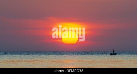 Pescatore solitario in una piccola barca a remi davanti a un tramonto luminoso, il mare calmo e vasto, la silhouette dell'isola di Koh Chang nella luce calda Foto Stock