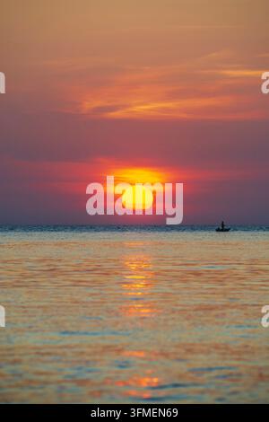 Pescatore solitario in una piccola barca a remi davanti a un tramonto luminoso, il mare calmo e vasto, la silhouette dell'isola di Koh Chang nella luce calda Foto Stock