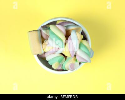 Marshmallow attorcigliati pastellati ammucchiati in una ciotola bianca su uno sfondo giallo brillante, dolcezza colorata flat lay per progetti di design di cibo e caramelle Foto Stock