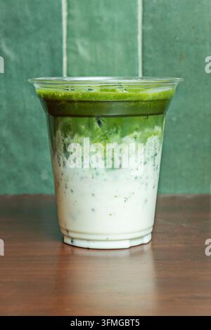 Latte freddo al matcha con strati di tè verde e latte in una tazza di plastica trasparente su uno sfondo di parete piastrellata verde. Foto Stock