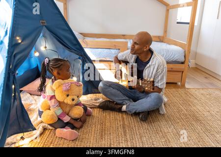 Padre e bambino afroamericani seduti su un tappeto da camera con teepee, suonando la chitarra, abbracciando il teddy. Famiglia, incollaggio, accogliente, soleggiato, tenda, peluche, orso Foto Stock