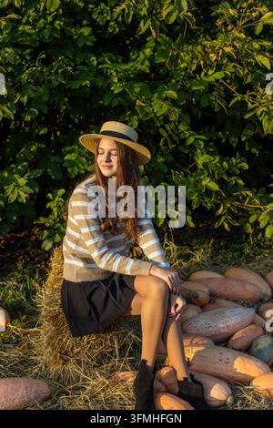 Giovane donna con cappello di paglia seduta su un ballotto di fieno accanto a zucche nella calda luce autunnale, ritratto stagionale tranquillo circondato da una vegetazione verde rigogliosa. Foto Stock
