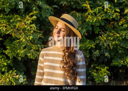 Giovane donna con cappello di paglia che gode della calda luce autunnale in un giardino verde, ritratto tranquillo all'aperto con lunghi capelli ricci e stagione naturale Foto Stock
