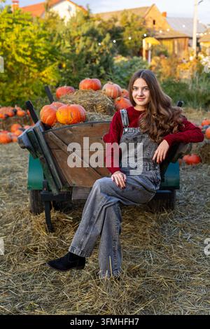 Giovane donna in tute jeans che posa accanto a un carretto rustico pieno di zucche e fieno, ritratto accogliente del raccolto autunnale alla calda luce della sera. Foto Stock