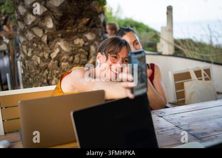 Coppia di giovani donne sorridenti che mostrano lavoro a distanza su telefono mobile con laptop in un bar caffè all'aperto su una terrazza, millennials felici trentenni con noma digitale Foto Stock