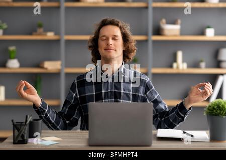 Giovane uomo d'affari che medita seduto con gli occhi chiusi, rilassandosi sul posto di lavoro in un ufficio moderno. Uomo che fa esercizio di rilassamento della meditazione al chiuso. Keep C Foto Stock