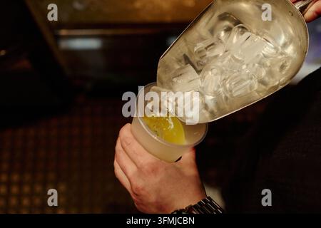 Barista che versa ghiaccio in una coppa da cocktail con un mestolo di metallo nel bar Foto Stock