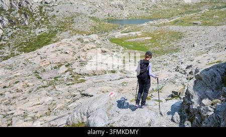 Vista posteriore di un'escursionista che scende un sentiero alpino impervio con bastoni da trekking e zaino, dirigendosi verso un piccolo lago di montagna attraverso un terreno roccioso ad alta quota nelle Alpi svizzere, Svizzera Foto Stock