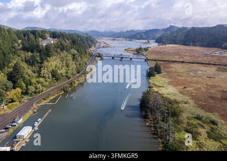 Vista aerea della linea ferroviaria di Coos Bay che attraversa il fiume Siuslaw vicino a Florence, Oregon, con barche da pesca al salmone, zone umide e un trasporto costiero c Foto Stock