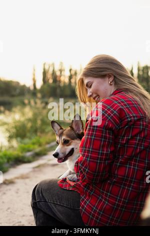 Giovane donna che cammina con il suo cane Pembroke Welsh Corgi in un parco verde in una giornata di sole. Donna e il suo animale domestico che si godono una passeggiata all'aperto circondati da alberi a Foto Stock