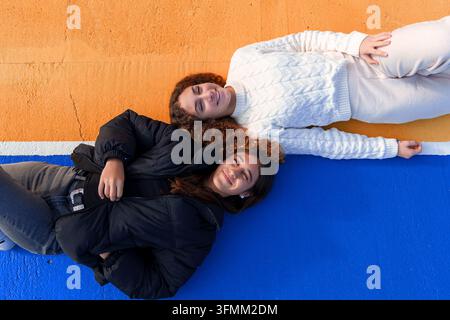 Due giovani amiche felici sdraiate su un pavimento colorato blu e arancione, sorridendo e guardando verso l'alto, esprimendo amicizia e gioia Foto Stock